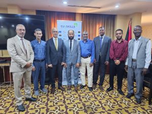 9F16F11B-8C3A-43DF-BB2E-D808C268E167 - جامعة سبها | Sebha University Group of eight men in business attire posing for a photo in a conference room with a EU4Skills banner in the background.