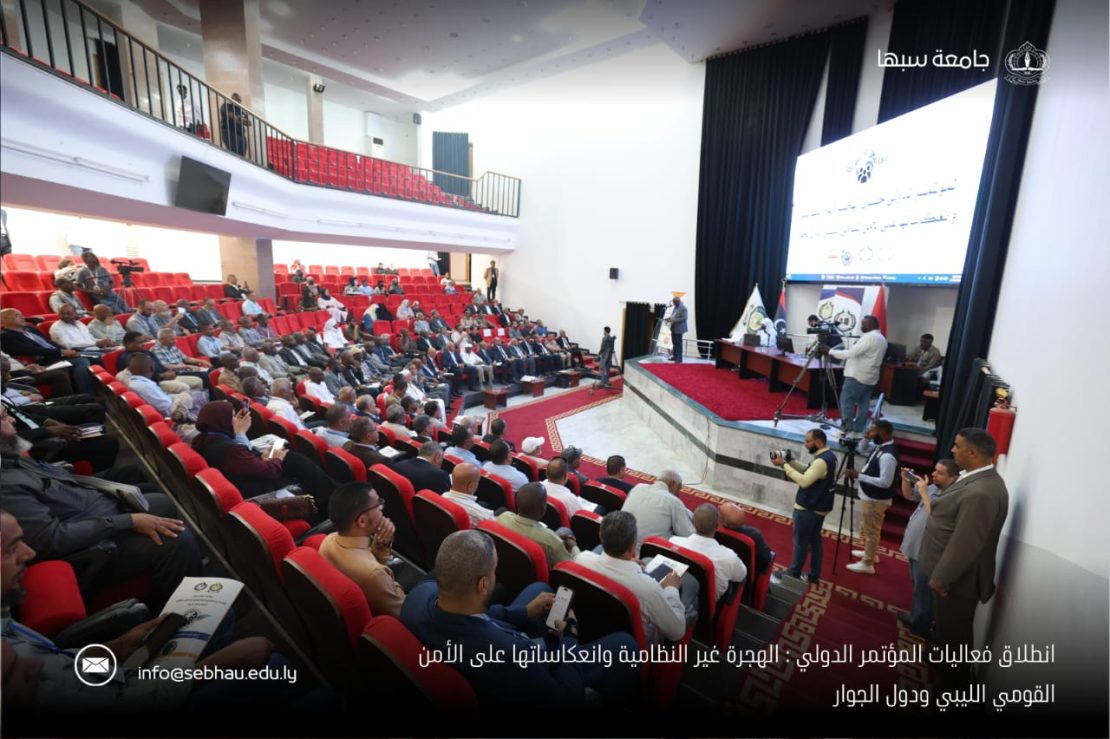 Audience seated in red chairs in a large auditorium, facing a stage with speakers, cameras, and a large projection screen.