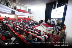 A7DEBCCE-8F68-4E9B-8968-2B050E83E980 - جامعة سبها | Sebha University Audience seated in red chairs in a large auditorium, facing a stage with speakers, cameras, and a large projection screen.