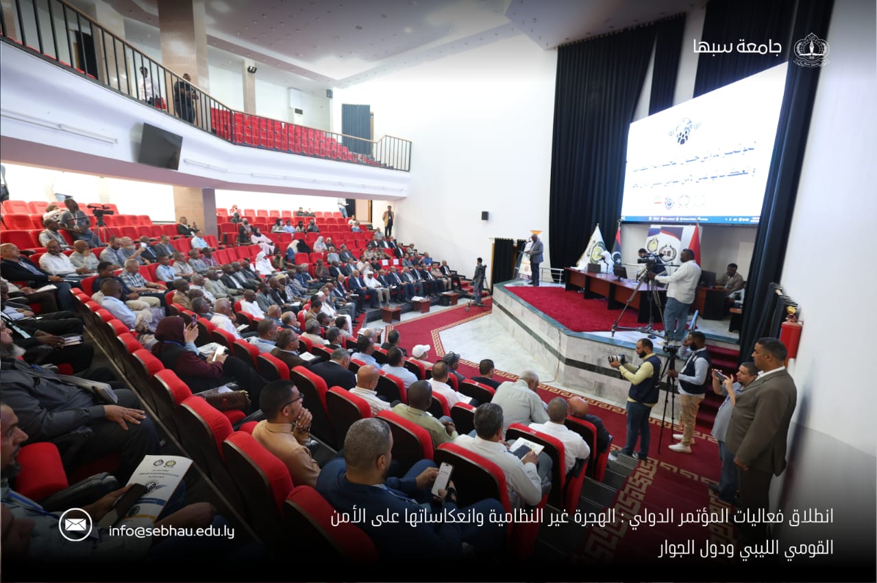 Audience seated in red chairs in a large auditorium, facing a stage with speakers, cameras, and a large projection screen.