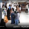 People at an indoor career fair with booths, banners, and a flower display; women in hijabs chat while photographers film in the background.