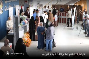 People at an indoor career fair with booths, banners, and a flower display; women in hijabs chat while photographers film in the background.