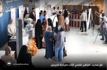 People at an indoor career fair with booths, banners, and a flower display; women in hijabs chat while photographers film in the background.