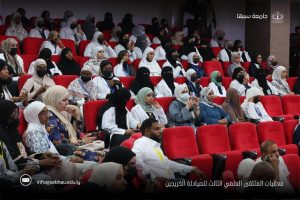 Audience in a conference hall with red theater seats; many women wearing hijabs and masks sit attentively.