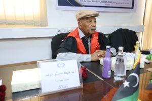 ABFBBBBD-1E27-4A85-88C9-7A5DFE4C23D3 - جامعة سبها | Sebha University Elderly man in a judge's robe and beige cap sits at a conference table during a formal meeting; flag and water bottles are on the desk.