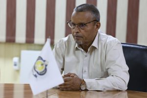Man in a light beige shirt wearing glasses sits at a conference table, hands folded, in an office with a flag on the desk behind him.