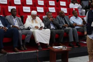 B076396B-9212-4902-902D-22D02F729863 - جامعة سبها | Sebha University Group of men in formal attire seated in red theater seats at a conference, one man in white traditional clothing looking at his phone.