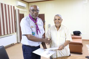 Two men shake hands while exchanging documents in a bright meeting room, one in a light uniform with a purple neckerchief and glasses.