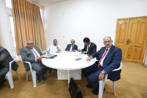 Five men in formal attire sit around a round white table in a meeting room, reviewing documents and discussing.