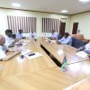 Group of people seated around a wooden conference table in a meeting room, taking notes and discussing a topic.