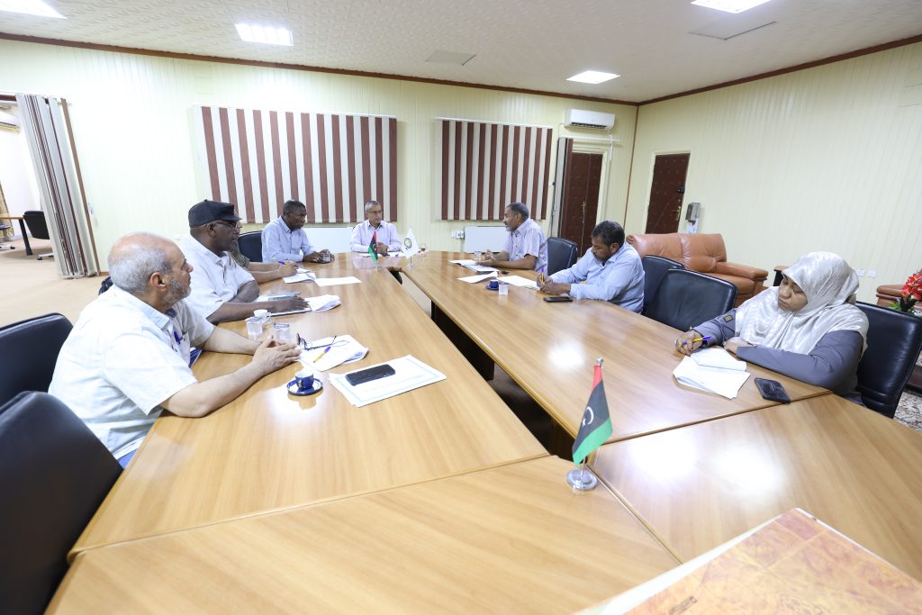 B52AC11C-EEE7-477B-858B-4AFB2051BDA6 - جامعة سبها | Sebha University Group of people seated around a wooden conference table in a meeting room, taking notes and discussing a topic.