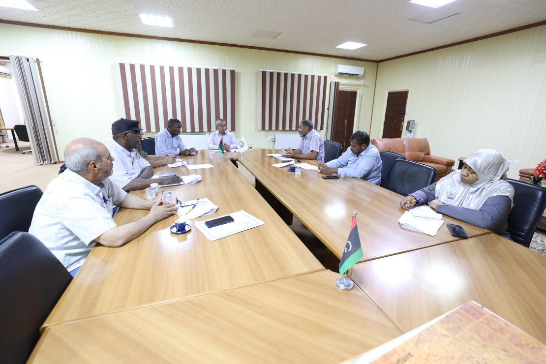 Group of people seated around a wooden conference table in a meeting room, taking notes and discussing a topic.
