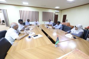 B52AC11C-EEE7-477B-858B-4AFB2051BDA6 - جامعة سبها | Sebha University Group of people seated around a wooden conference table in a meeting room, taking notes and discussing a topic.