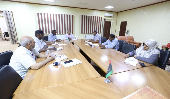 Group of people seated around a wooden conference table in a meeting room, taking notes and discussing a topic.