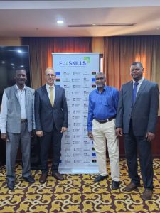 B58C8D58-CCDF-418C-BA7F-72728DDC8D01 - جامعة سبها | Sebha University Four men in business attire pose for a photo in front of a EU4Skills banner at a formal event in a conference room.