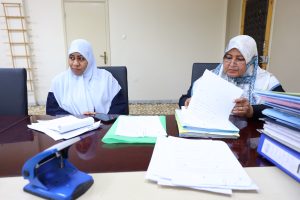 Two women in hijabs sit at a meeting table covered with papers, looking focused