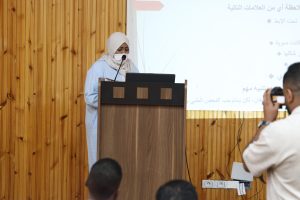 Woman wearing a beige hijab and mask speaks at a wooden podium with a laptop and microphone, in front of a projection screen.