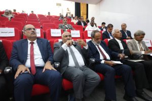 B896866A-4C37-4DFF-9ACA-B03534C466C7 - جامعة سبها | Sebha University Group of men in suits seated in red auditorium chairs at a formal conference; front row focused on speakers and discussion