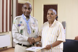 Two men shake hands, one in a beige uniform with badges and a blue scarf, the other in a striped shirt, holding a certificate.