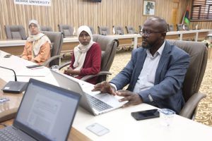 BC5FB305-A6C7-43F4-B8B9-74CADFD27200 - جامعة سبها | Sebha University Three professionals sit at a long conference table with laptops in a wood-paneled meeting room, a banner visible in the background.