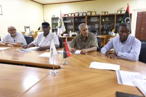 BEC18547-7460-4AA5-91D2-33F81C9B14A2 - جامعة سبها | Sebha University Four men sit at a long conference table in a wood-paneled room, examining documents with flags visible on the table behind them.