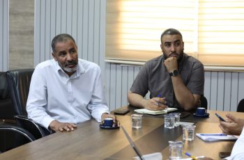 Two men sit at a conference table in a meeting room, one in a white shirt speaking while the other in a gray shirt listens with a pen and notebook nearby.