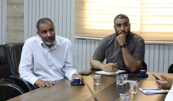 BF14BEBB-9966-4F36-9A97-991C3A40F830 - جامعة سبها | Sebha University Two men sit at a conference table in a meeting room, one in a white shirt speaking while the other in a gray shirt listens with a pen and notebook nearby.