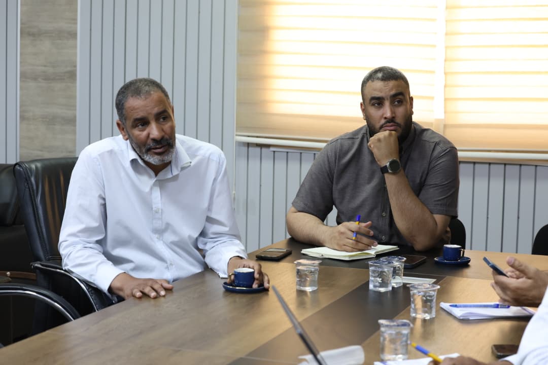 Two men sit at a conference table in a meeting room, one in a white shirt speaking while the other in a gray shirt listens with a pen and notebook nearby.