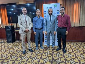 BFEABFE0-E06F-4C0D-88A7-F7E3F731BF1B - جامعة سبها | Sebha University Four men in business attire pose for a photo at an EU4Skills event, standing in front of a banner and a large screen.