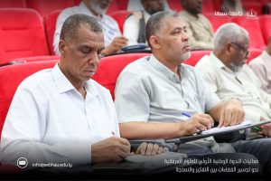 Audience of men seated in a red auditorium, taking notes during a conference, with an Arabic university logo visible in the top right corner.