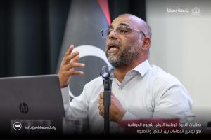 Speaker wearing glasses and a white shirt speaks into a microphone at a conference; laptop and Arabic logos visible in the background.