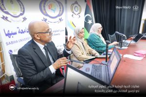 Man in a suit speaks into a handheld microphone at a panel discussion, with two women in hijabs and laptops on the table behind him.