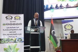 Man in a white shirt and black vest speaks at a podium during a conference, with banners and a Libyan flag nearby.