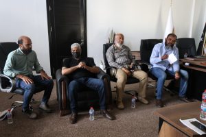C7AE5FB9-09F6-4292-AC9F-1BFC9D0DE9F5 - جامعة سبها | Sebha University Four men sit in an office coming a meeting, wearing masks, with water bottles on the floor nearby and a desk on the right.