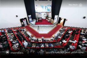 Lecture hall during a conference with a panel on stage, audience in red seats, banners and a projection screen behind the panelists, Libyan flag visible on stage.