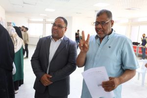 Two men at an indoor event: one in a dark suit and white shirt with hands clasped, the other in a light blue shirt holding papers and flashing a peace sign while facing the camera.