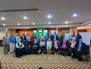 CE22D997-817F-4EC0-83BC-88B54471DE8C - جامعة سبها | Sebha University Group of professionals posing for a conference room group photo, with banners and a screen in the background.