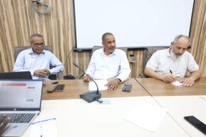 CE2C422E-638E-411B-AFD6-CCE0D1DB8E3A - جامعة سبها | Sebha University Three men sit at a conference table in a meeting room, one typing on a laptop, two taking notes, with a projector screen behind them.