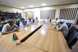 Group of men in uniforms seated around a long conference table in a meeting room, with Libyan flags on the table and a flag in the background.