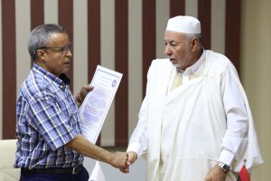 DA039D1C-4F16-4E23-BEC6-AE68CEB998F9 - جامعة سبها | Sebha University Two men shake hands as one presents a document or certificate during a formal ceremony.