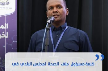Man speaks at a podium during a conference, wearing a dark blue shirt and a blue lanyard with a microphone in front of a banner backdrop.