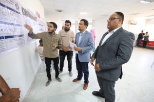 Group of men examining large posters with Arabic text on a wall in a bright hallway; one man points and explains to the others.