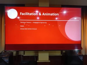 DD641EC4-BCB0-4C23-8F52-E00D6023AC32 - جامعة سبها | Sebha University Large presentation slide on a red background reading 'Facilitation & Animation' with trainer names listed beneath; a training workshop title screen on a big monitor.