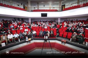 Audience seated in a large auditorium with red cushioned seats, a front‑stage area with cameras on tripods, and an Arabic caption about the third scientific forum.