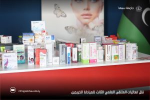 Row of assorted medicine boxes and vitamins lined up on a white counter at a health expo backdrop, with a large poster and Arabic text in the background.] ,