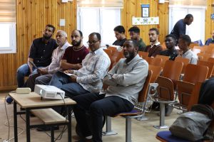 Group of men seated in a wood-paneled classroom, attentively listening with a projector on the front table nearby.