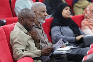 E3331FC3-A748-4BA7-84FB-6FCA72641130 - جامعة سبها | Sebha University Man in a beige jacket reads a program while seated in red theater seats at an event, with other attendees nearby.