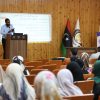 Lecturer at a podium presenting to an audience in a wooden-floored lecture hall with flags and a projection screen displaying Arabic text.