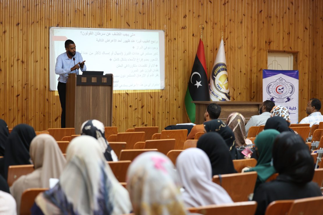 Lecturer at a podium presenting to an audience in a wooden-floored lecture hall with flags and a projection screen displaying Arabic text.