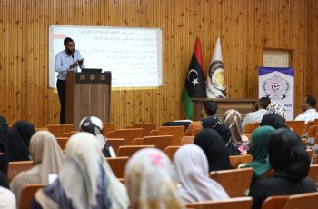Lecturer at a podium presenting to an audience in a wooden-floored lecture hall with flags and a projection screen displaying Arabic text.